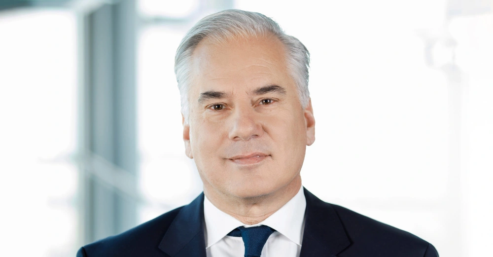 Mann lächelt leicht stehend Umgebung ist ein helles Büro mit unscharfem Hintergrund. Keine sichtbaren Texte.