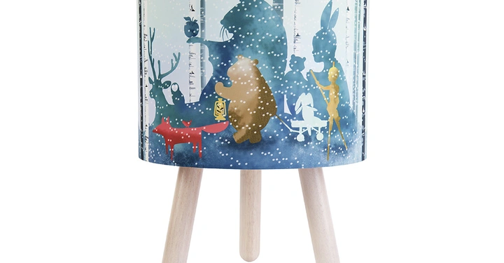Galerie-Bild 7: Eine runde Tischlampe steht auf drei Holzbeinen ein Schirm mit Illustrationen von Tieren wie Bären Füchsen Rehen und Hasen in einem verschneiten Wald.