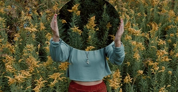 Person hält einen runden Spiegel im Feld mit gelben Blumen der Spiegel reflektiert die Umgebung wodurch das Gesicht der Person unsichtbar wird die Person trägt ein türkises Oberteil und eine rote Hose