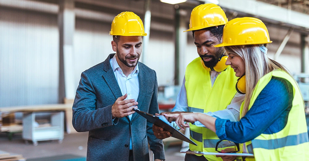 Eine Frau und zwei Männer in gelben Schutzhelmen und blauen Schutzwesten inspizieren eine Baustelle und unterhalten sich entspannt.