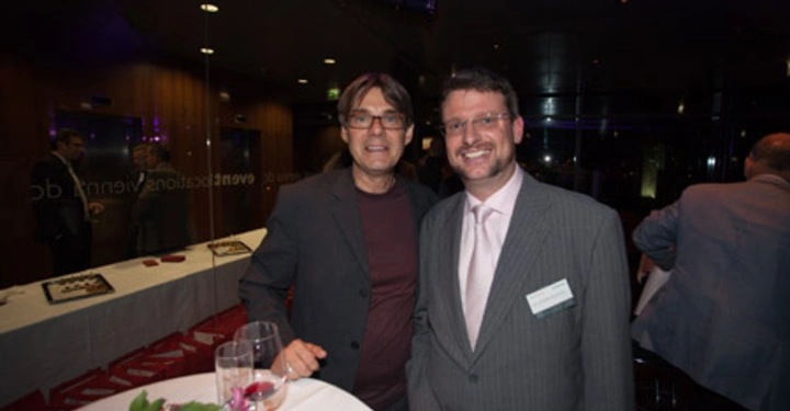 Galerie-Bild 74: Zwei Männer stehend lächelnd in einem modernen Indoor-Veranstaltungsraum einer trägt einen Anzug der andere ein schwarzes Hemd und eine Brille sie stehen an einem Tisch mit Getränken und Blumen