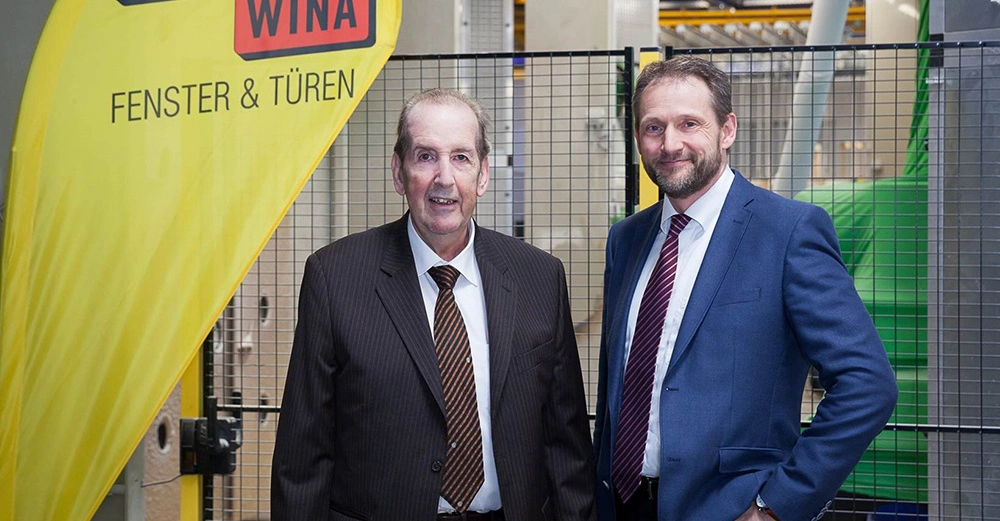 Zwei Männer in Anzügen stehen nebeneinander, lächelnd in einer modernen Fabrikumgebung. Links ein gelbes Schild mit der Aufschrift WINA FENSTER & TÜREN. Im Hintergrund Maschinerie und Metallgitter.