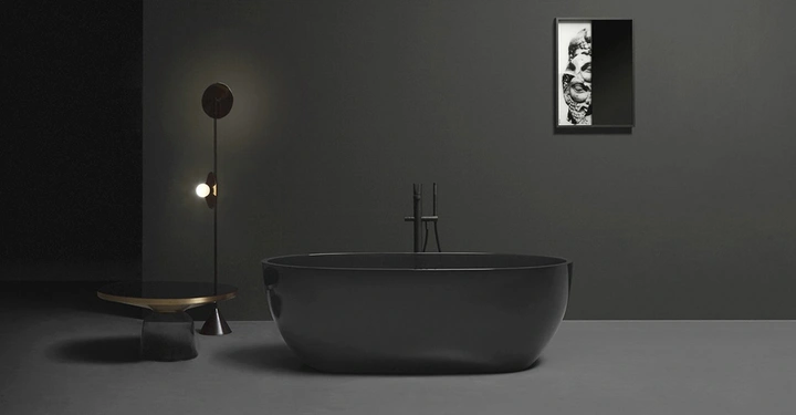 Freistehende schwarze Badewanne steht vor einer dunklen Wand neben einer modernen Lampe und einem kleinen Tisch, Bild an der Wand zeigt Gesichterrelief.