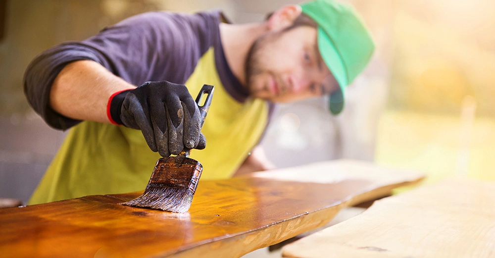 Person streicht Holzbrett mit Pinsel trägt grüne Mütze und Handschuhe befindet sich in heller Werkstattumgebung