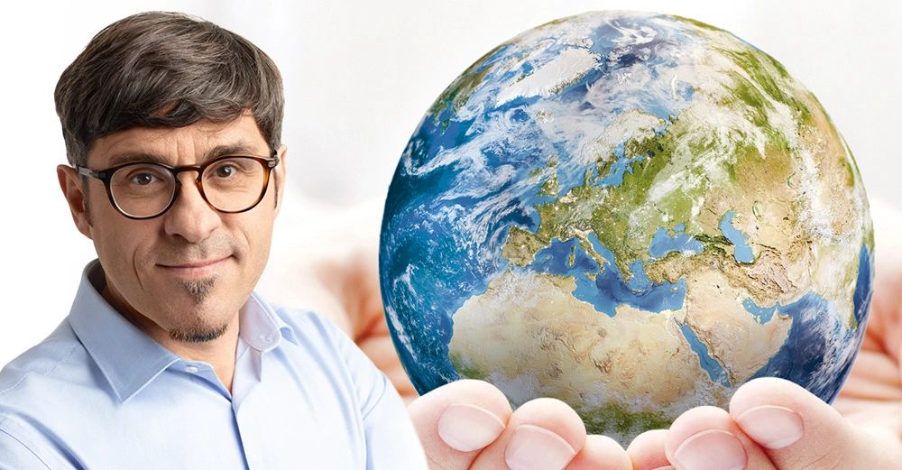 Mann mit Brille neben einer großen Weltkugel die von Händen behutsam gehalten wird vor einem hellen Hintergrund.