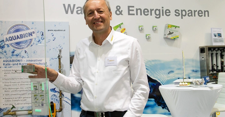 Galerie-Bild 9: Mann hält Gerät vor sich und lächelt energetisch in die Kamera in einer Ausstellungshalle. Im Hintergrund befinden sich Produkte und Werbeschilder mit Texten "Wasseraufbereitung", "Wasser & Energie sparen", "AQUABION", "innovative Kalk- und Korrosionsschutz", und "Nachhaltigkeit".