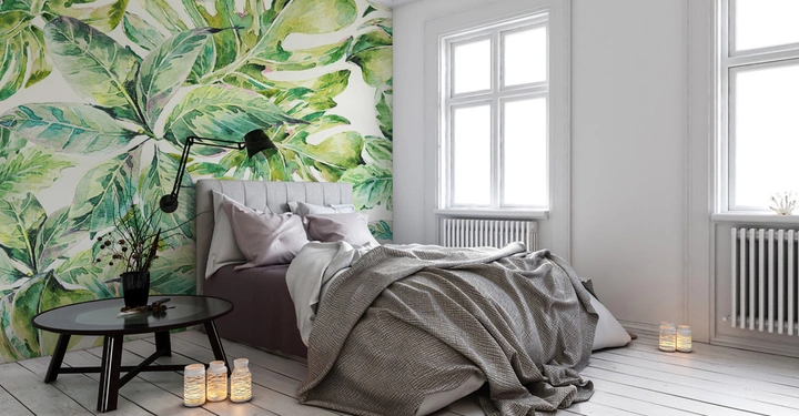 Galerie-Bild 6: Ein Bett steht in einem Zimmer mit einem großen Blattmuster an der Wand ungemachtes Bett mit grauer Decke und Kissen daneben ein runder Tisch Kerzenlichter auf dem Boden helle Fenster und Holzboden.