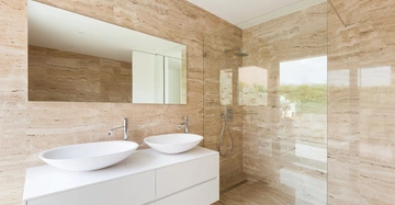 Doppelwaschbecken auf weißem Schrank reflektiert in großem Spiegel daneben begehbare Dusche mit Glastrennwand helle Steinwände und Boden in modernem Badezimmerambiente.