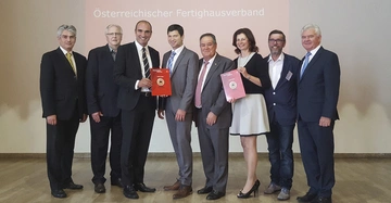 Acht Personen stehen nebeneinander und halten Preiszertifikate lächelnd, während sie in einem Veranstaltungsraum posieren. Hintergrundtext: "57. Ordentliche Generalversammlung Österreichischer Fertighausverband".