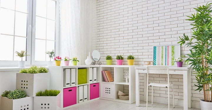 Weiße Regale mit bunten Blumentöpfen und Ordnern stehen vor einer weiß gestrichenen Ziegelwand neben einem mit Pflanzen dekorierten Schreibtisch in einem hellen Raum mit großen Fenstern und Vorhängen