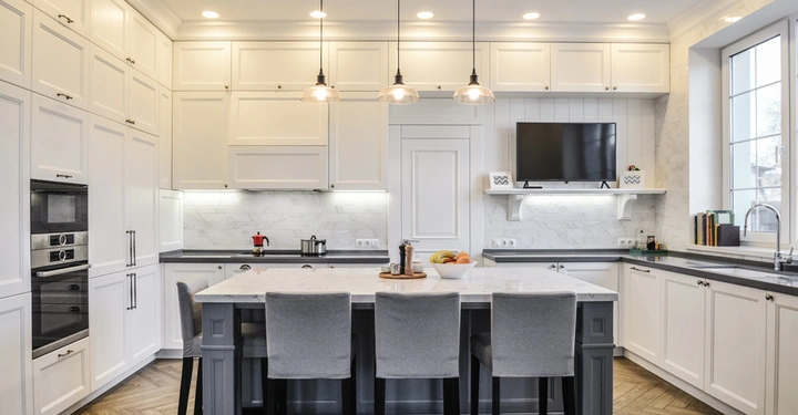 Küchenschrank hängen an der Wand über Arbeitsplatten Kücheninsel mit drei Stühlen und Obstschale moderne Geräte und Pendelleuchten in einem hell beleuchteten Raum mit großen Fenster.