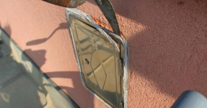 Galerie-Bild 7: Verrostete Kelle hebt eine Metallplatte von einer rosa Wand ab. Auf der rechten Seite ist ein blaues Rohr sichtbar. Schatten fallen auf die Wand und den Boden.