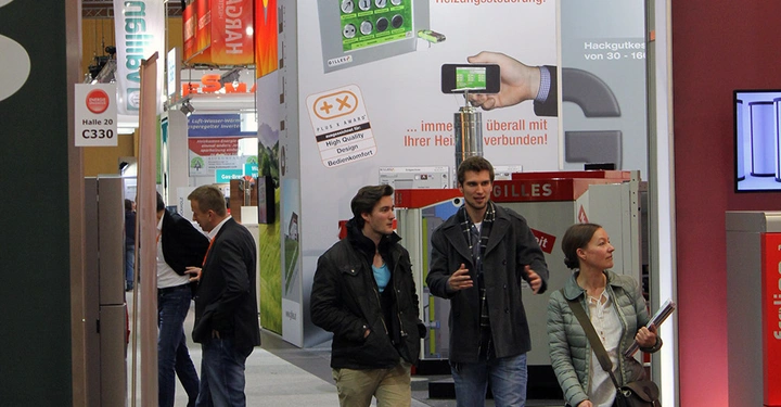 Galerie-Bild 7: Drei Personen gehen durch eine Messehalle umgeben von farbenfrohen Werbebannern und Ständen. Texte: Halle 20 C330; intelligentes Heizungssteuerung; immer überall mit Ihrer Heizung verbunden; High Quality Design Bedienkomfort.