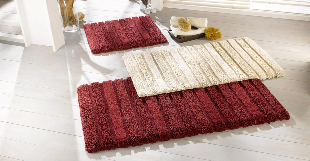 Drei Badezimmerteppiche liegen auf einem hellen Holzboden. Zwei sind rot und gestreift, einer ist weiß und gestreift. Im Hintergrund befinden sich Handtücher und Bürsten.
