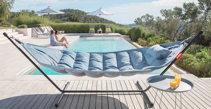 Galerie-Bild 4: Eine blaue Hängematte hängt auf einer Terrasse neben einem Pool. Zwei Personen sitzen am Poolrand. Die Umgebung ist grün und hügelig. Ein kleiner Tisch steht neben der Hängematte.