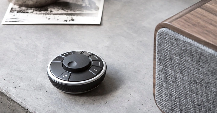 Ein runder schwarzer Fernbedienungsregler mit mehreren Tasten liegt auf einer grauen Betonoberfläche neben einem hölzernen Lautsprecher. Im Hintergrund ist ein Schwarzweißfoto teilweise sichtbar. Tastenbeschriftungen: BACK MENU SOURCE 1+ FAV SET POWER