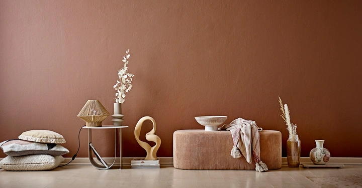 Stapel Kissen und Tischlampe auf einem Beistelltisch in einem Wohnzimmer neben einer Bank mit Decke Vase und abstrakter Skulptur vor brauner Wand.