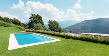 Ein rechteckiger Swimmingpool liegt in einem gepflegten grünen Rasen umgeben von blühenden Sträuchern Berge und blauer Himmel mit Wolken im Hintergrund