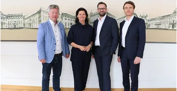 Drei Männer und eine Frau stehen vor einer großen Wand mit Gebäuden auf großer Leinwand.