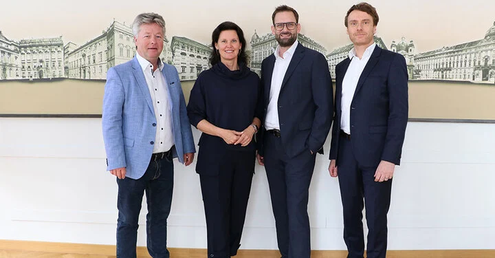 Drei Männer und eine Frau stehen vor einer großen Wand mit Gebäuden auf großer Leinwand.