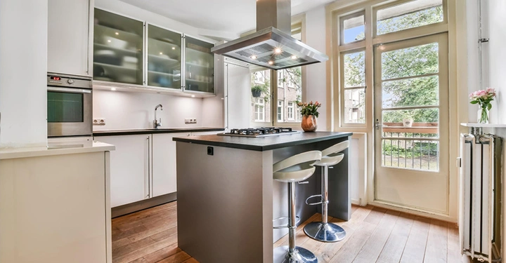 moderne Holzküche mit grauer Kücheninsel, Dunstabzugshaube und Gasherd. Rechts große helle Fensterfront.