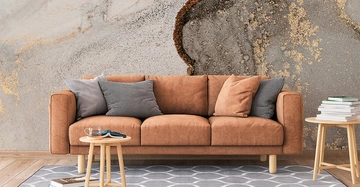 Braunes Sofa steht vor einer künstlerisch gestalteten Wand im Muster eines Marmors. Kissen liegen auf dem Sofa. Ein kleiner runder Holztisch mit Büchern und einem Glas steht davor.