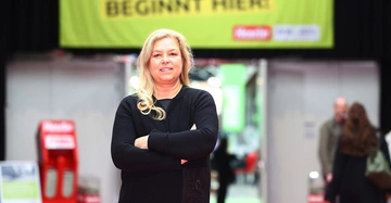 Frau mit langen blonden Haaren in schwarzem Outfit steht mit verschränkten Armen vor einer Leinwand mit dem Logo der Messe KlimaZukunft Österreich.