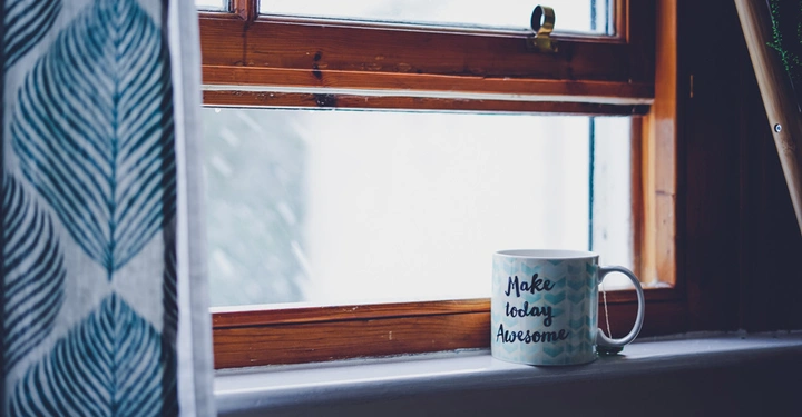 Eine Tasse mit der Aufschrift Make today Awesome steht auf einer Fensterbank aus Holz mit Blick auf eine verschneite Landschaft. Ein Vorhang mit Blattmuster hängt daneben.