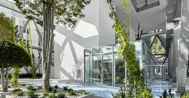 Ein moderner Glaspavillon steht inmitten eines grünen Innenhofs mit Bäumen und Sträuchern umgeben von hohen weißen Gebäuden mit geometrischen Schattierungen auf den Wänden.