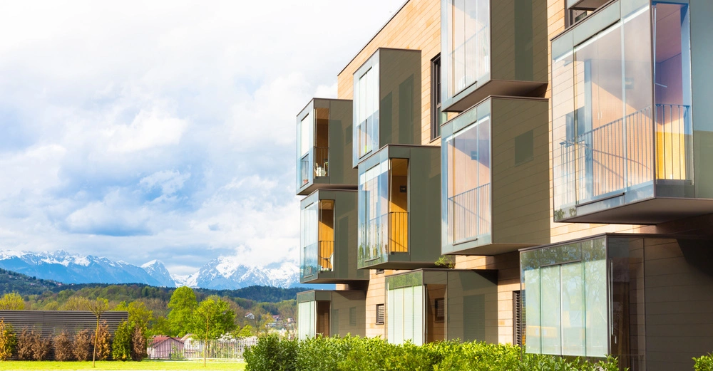 Moderne Wohnungen mit großen Glasfenstern und Balkonen, die in die Luft ragen. Sie befinden sich in einer ruhigen Umgebung mit grünen Büschen im Vordergrund und schneebedeckten Bergen im Hintergrund.