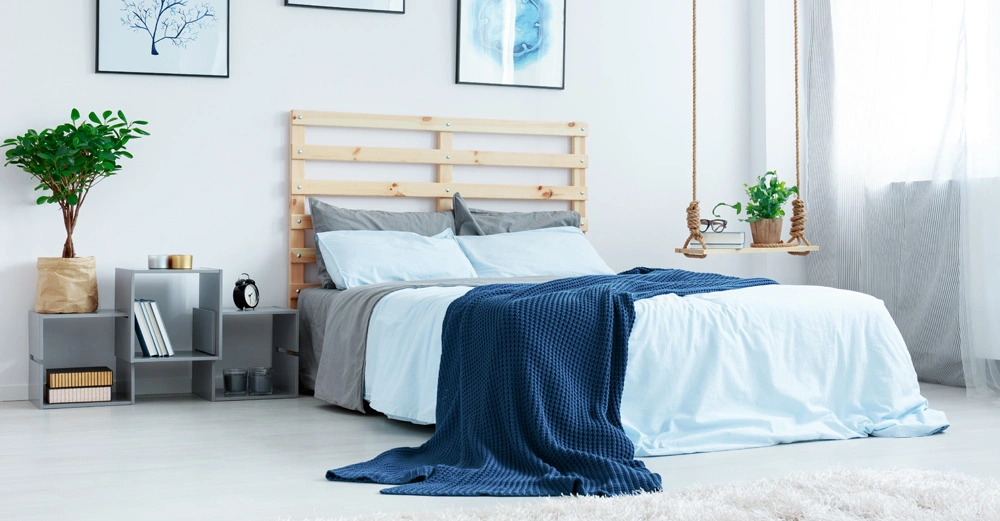 Bett mit hellblauer Bettwäsche und dunkelblauer Decke steht vor einer Holzkopfteilwand mit drei Bildern. Links ein Nachttisch und Pflanze, rechts hängendes Regal, Vorhänge und Fenster.