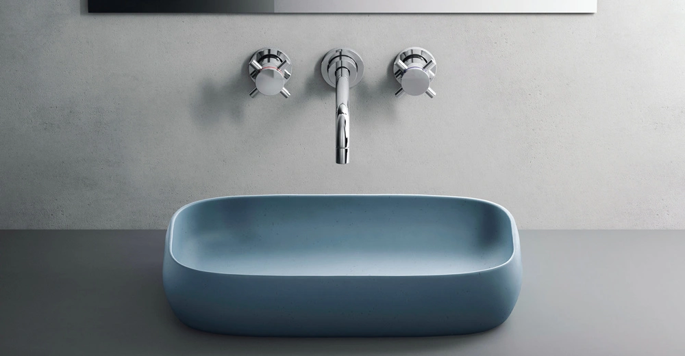 Ein rechteckiges graues Waschbecken steht auf einer grauen Oberfläche mit einer silbernen Wandarmatur darüber in einem minimalistischen Badezimmer.