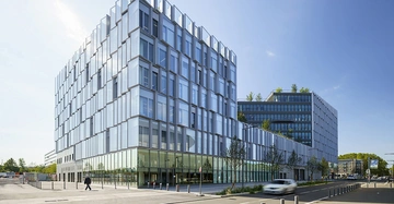 Großes modernes Glasgebäude links Menschen gehend Eingang passierend; rechts Bäume Pflanzen auf alten Dächern in städtischer Umgebung.