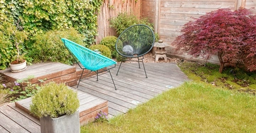 Zwei farbige Stühle stehen auf einer Holzterrasse in einem gut gepflegten Garten, umgeben von Pflanzen, Holzlattenzäunen und einem kleinen Ziersteinlaterne.