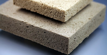 Zwei rechteckige Schwämme liegen übereinander auf einer glatten, grauen Oberfläche. Sie haben eine raue Textur und sind in verschiedenen Brauntönen.