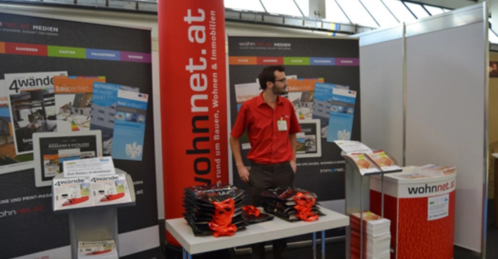 Galerie-Bild 2: Mann steht hinter Tisch mit Stapeln von Beuteln und Broschüren in Messestand mit wohnnet.at-Werbung umgeben von Broschürenständen und Bannern in einer Messehalle. Text: wohnnet.at Ihr Partner rund um Bauen Wohnen und Immobilien.