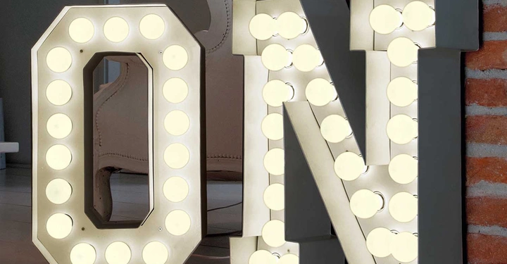 Große beleuchtete Buchstaben D und N stehen nebeneinander. Der Hintergrund zeigt eine Ziegelwand und ein modernes Zimmer mit einem Stuhl im Hintergrund.