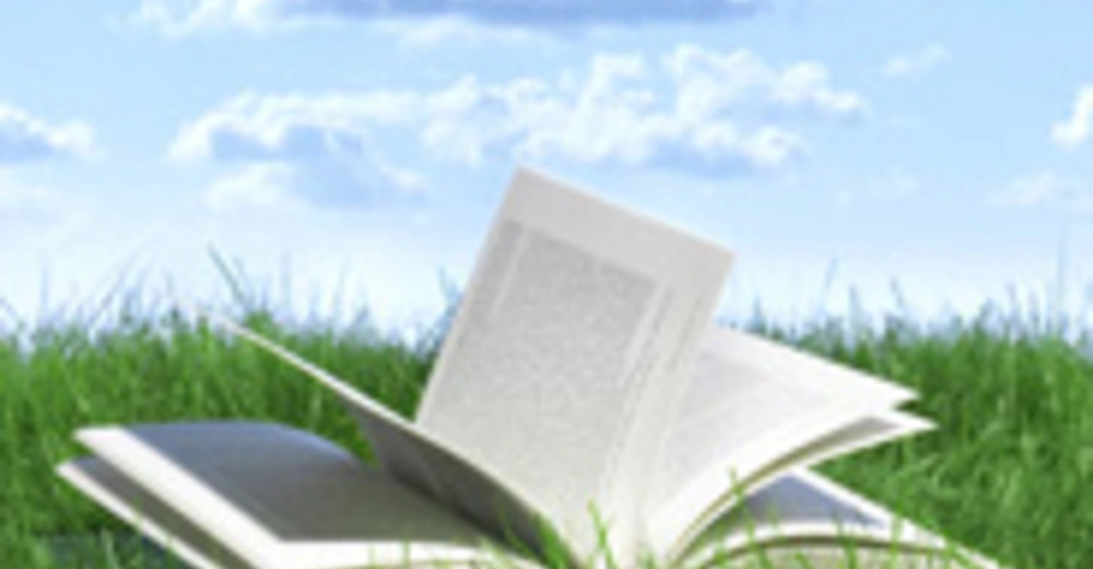 Ein geöffnetes Buch liegt im Gras bei Tageslicht umgeben von blauen Himmel und weißen Wolken