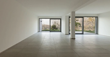 Ein großer leerer Raum mit grauen Fliesenboden und weißen Wänden hat an der Rückseite mehrere große Glas-Schiebetüren, die zu einem Garten und Bäumen im Freien führen.