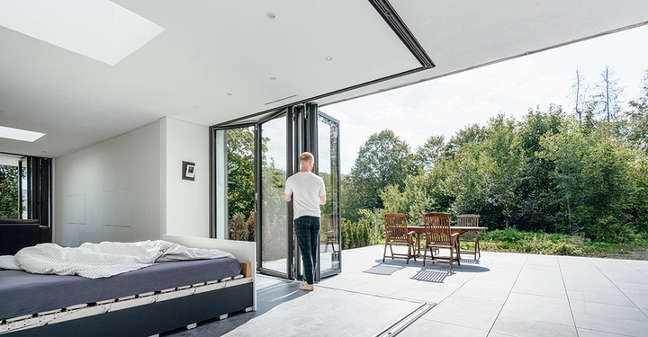 Galerie-Bild 3: Eine Person öffnet Glastüren in einem modernen Schlafzimmer mit ungemachtem Bett das ins Freie führt wo Möbel auf einer Terrasse stehen umgeben von grünen Bäumen und blauem Himmel