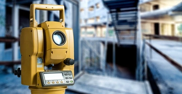 Gelbes Vermessungsgerät mit Display und Tasten steht still in einem unfertigen Gebäude mit Betontreppen und Gerüsten