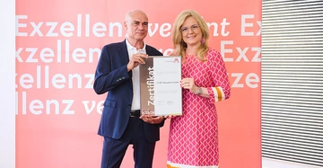 Zwei Personen halten ein Zertifikat vor einem Hintergrund mit dem Text Exzellenz. Das Zertifikat lautet Leittechnik Exzellenz Zertifikat GLOBRT Bussysteme GmbH 2023 24.