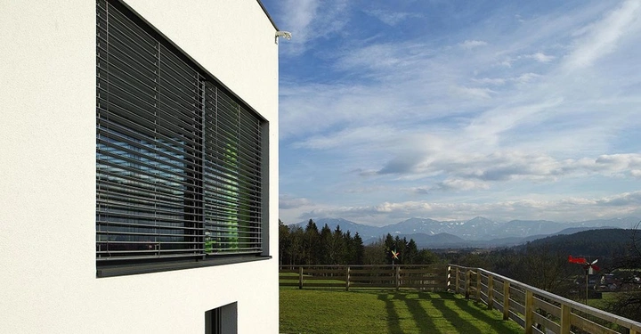 Galerie-Bild 1: angeschnittene hellbeige Fassade mit großem Fenster und dunklen Rollläden und großer Garten mit Holzzaun, blauer Himmel und Wolken
