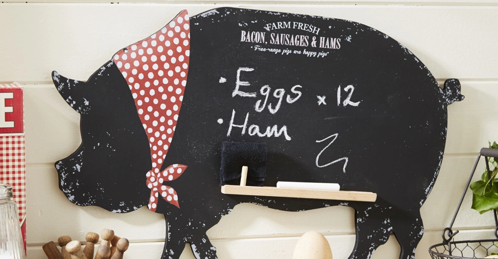 Schweineförmige Tafel mit rot-weiß gepunktetem Kopftuch listet Eier x 12 und Schinken auf im Küchenbereich mit Gewürzgläsern und Dekorationsgegenständen drumherum. Text oben: FARM FRESH BACON SAUSAGES & HAMS Free-range pigs are happy pigs