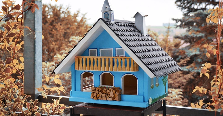 Galerie-Bild 4: Ein blaues, hölzernes Vogelhaus steht still auf einem Tisch inmitten von herbstlichen Blättern und umgebenden Bäumen.