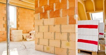 Unvollendete Innenwand aus Ziegelsteinen steht in einem rohbauartigen Raum mit sichtbaren Holzbalken Decke. Stapel von Baumaterialien und Isolierungspaketen sind im Hintergrund verteilt. Kein Text vorhanden.