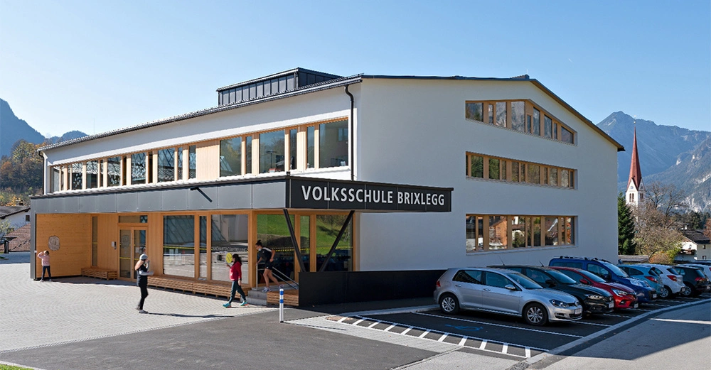 Gebäude der Volksschule Brixlegg Menschen gehen hinein im Vordergrund parken Autos im Kontext von Alpengipfeln und einem Kirchturm im Hintergrund