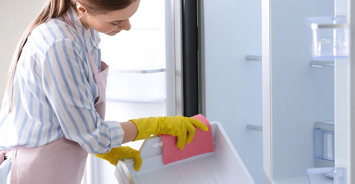 Eine Frau reinigt mit einem rosa Tuch und gelben Gummihandschuhen den Innenraum eines offenen Kühlschranks in einer häuslichen Küche.