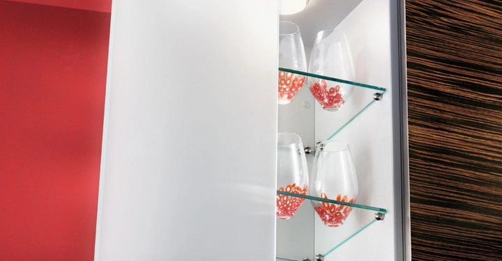 Galerie-Bild 5: Holzschrank mit geöffnetem Türchen stellt dekorative Gläser und rote Teller auf Glasregalen aus in einem Raum mit roten Wänden und weißer Decke.