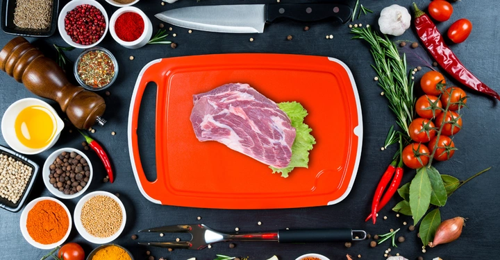 Ein Stück rohes Fleisch liegt auf einem roten Schneidebrett in der Mitte etlicher Gewürze Zutaten und Küchenutensilien auf einem dunklen Hintergrund verteilt.